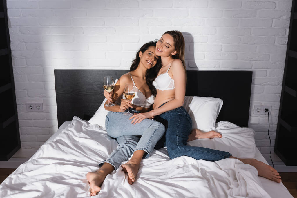 happy lesbian couple in bras holding glasses of white wine while resting on bed - Fotografie, Obrázek