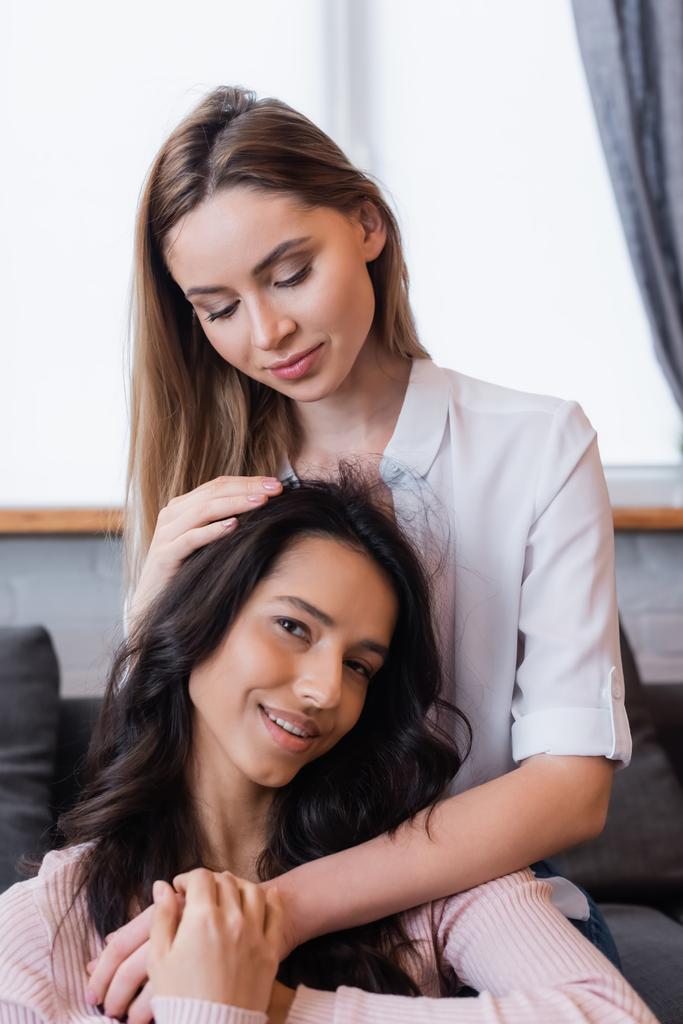 smiling lesbian woman embracing brunette girlfriend in living room - Photo, Image