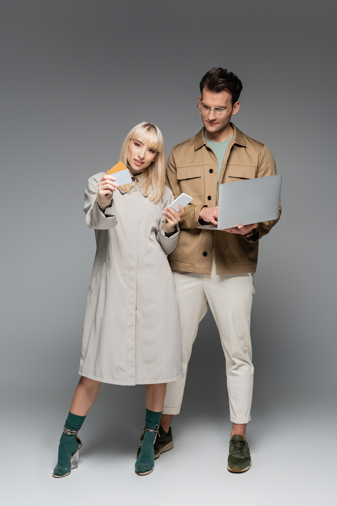 young woman in shirt dress holding smartphone and credit cards near stylish man using laptop on grey - Photo, Image