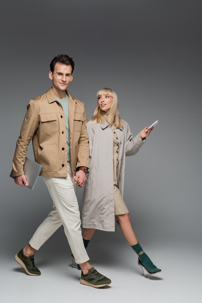 full length of trendy couple holding hands while walking with gadgets on grey - Photo, Image