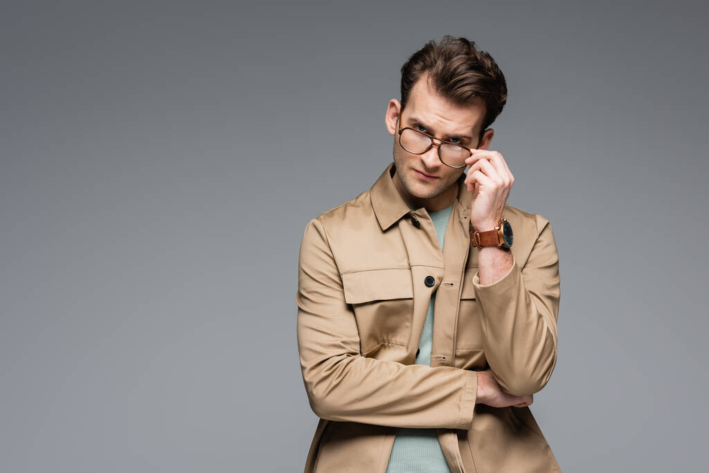 модний чоловік в куртці регулюючи окуляри, позує ізольовано на сірому
 - Фото, зображення
