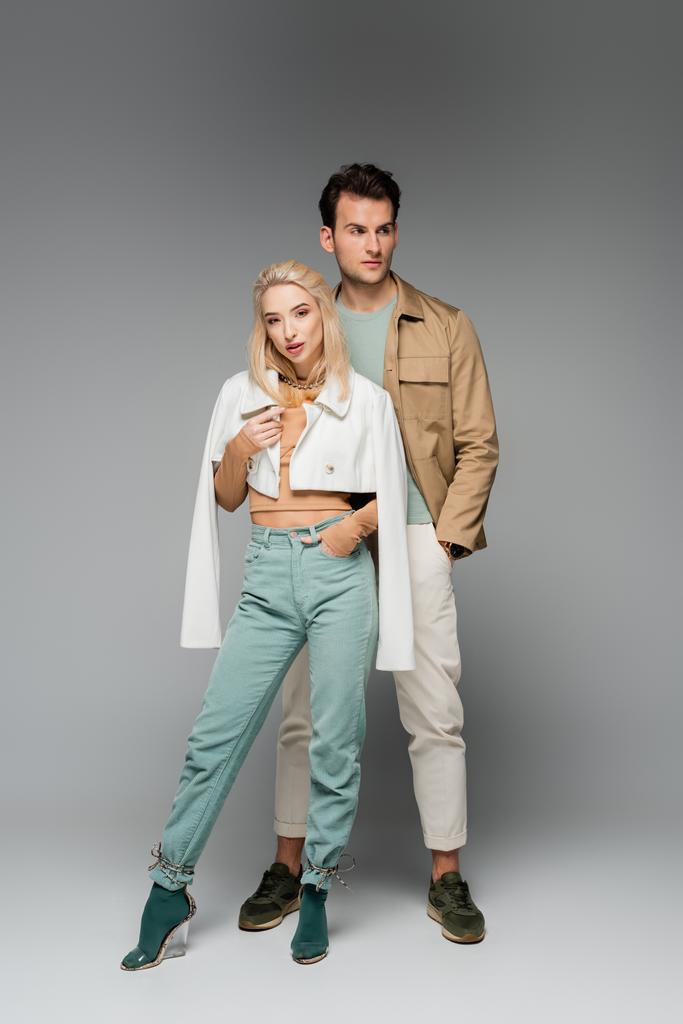 volledige lengte van trendy modellen in broeken en jassen poseren met de handen in zakken op grijs - Foto, afbeelding