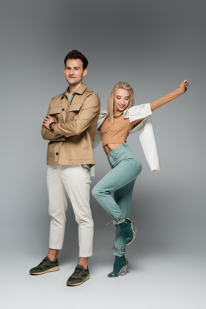 full length of stylish models in pants and jackets posing and smiling on grey - Photo, Image