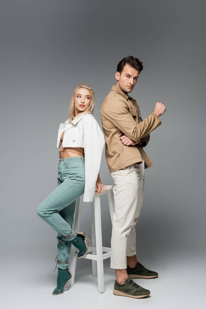 longueur totale de jeunes modèles élégants en pantalon et vestes posant près de chaise blanche sur gris - Photo, image