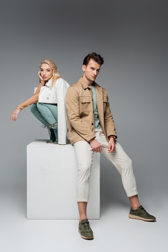 full length of stylish models in pants and jackets posing while sitting on white cube on grey - Photo, Image