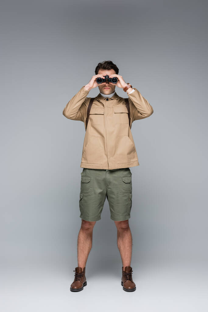 volledige lengte van trendy reiziger in shorts en jas kijkend door verrekijker op grijs  - Foto, afbeelding