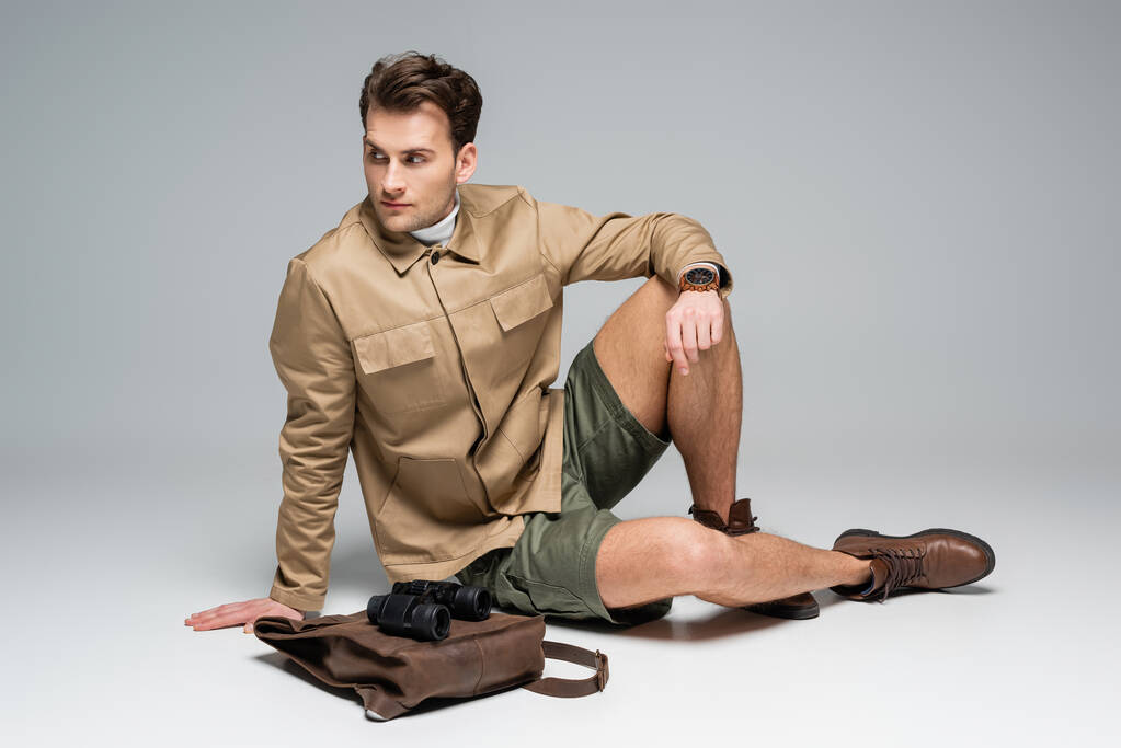 longitud completa de viajero de moda en pantalones cortos y chaqueta sentado cerca de la mochila de cuero con binoculares en gris  - Foto, imagen