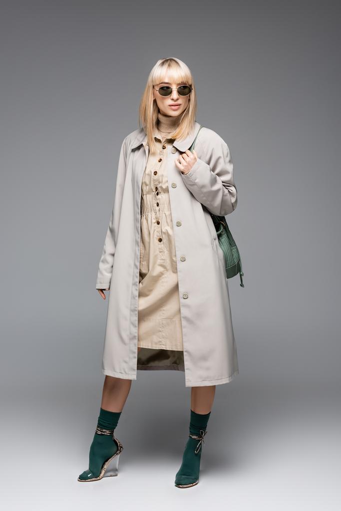 full length of fashionable woman in sunglasses and trench coat posing with green bag on grey - Photo, Image