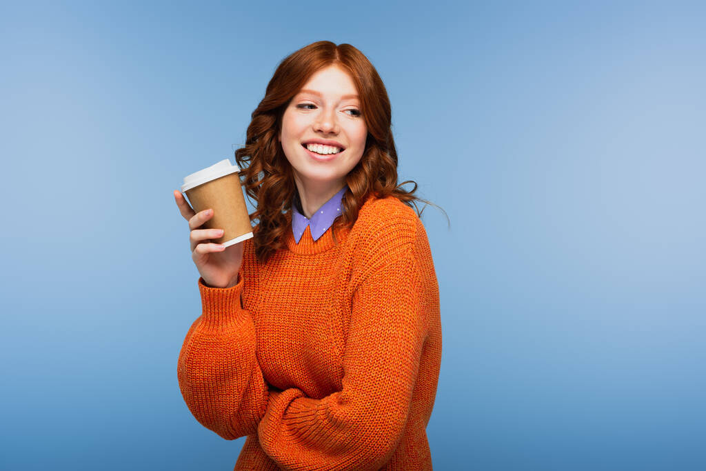positive redhead woman in orange sweater holding coffee to go isolated on blue - Photo, Image