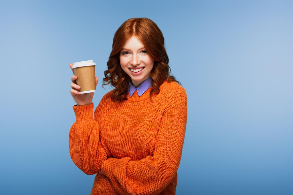happy redhead woman in orange sweater holding coffee to go isolated on blue - Photo, Image