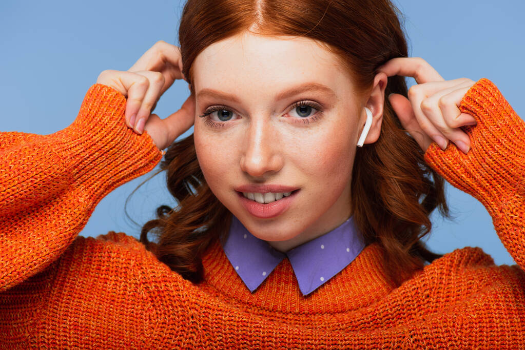 mulher ruiva feliz ouvindo música em fones de ouvido sem fio e ajustando o cabelo isolado no azul - Foto, Imagem