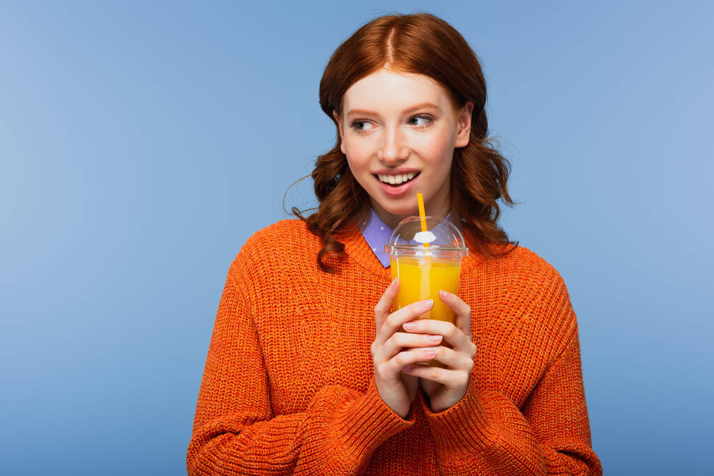 χαμογελαστή κοκκινομάλλα γυναίκα σε πουλόβερ κρατώντας φρέσκο χυμό πορτοκαλιού σε πλαστικό κύπελλο απομονώνονται σε μπλε - Φωτογραφία, εικόνα