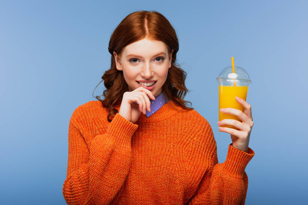 mujer pelirroja positiva en suéter que contiene jugo de naranja fresco en taza de plástico aislado en azul - Foto, imagen