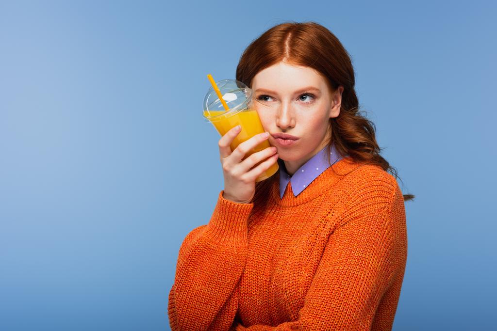 Скучная рыжая женщина в свитере держит свежий апельсиновый сок в пластиковой чашке изолированы на синий - Фото, изображение