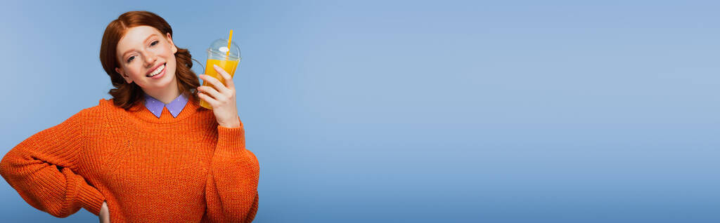 mujer pelirroja feliz en suéter que contiene jugo de naranja fresco en taza de plástico aislado en azul, pancarta - Foto, Imagen