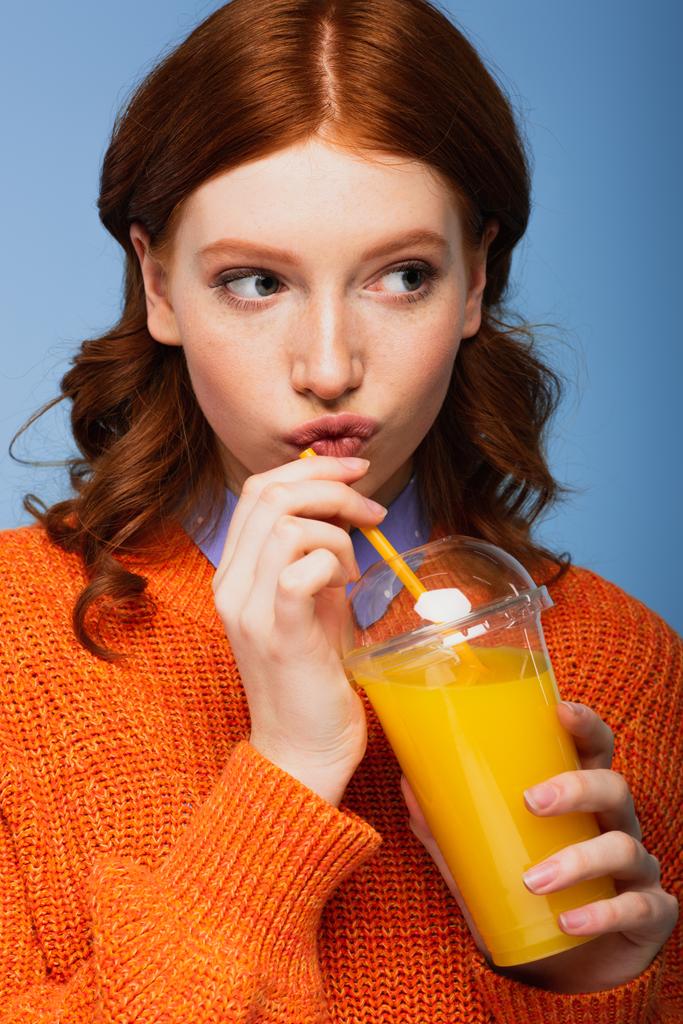 redhead woman in sweater pouting lips and holding fresh orange juice in plastic cup isolated on blue - Photo, Image