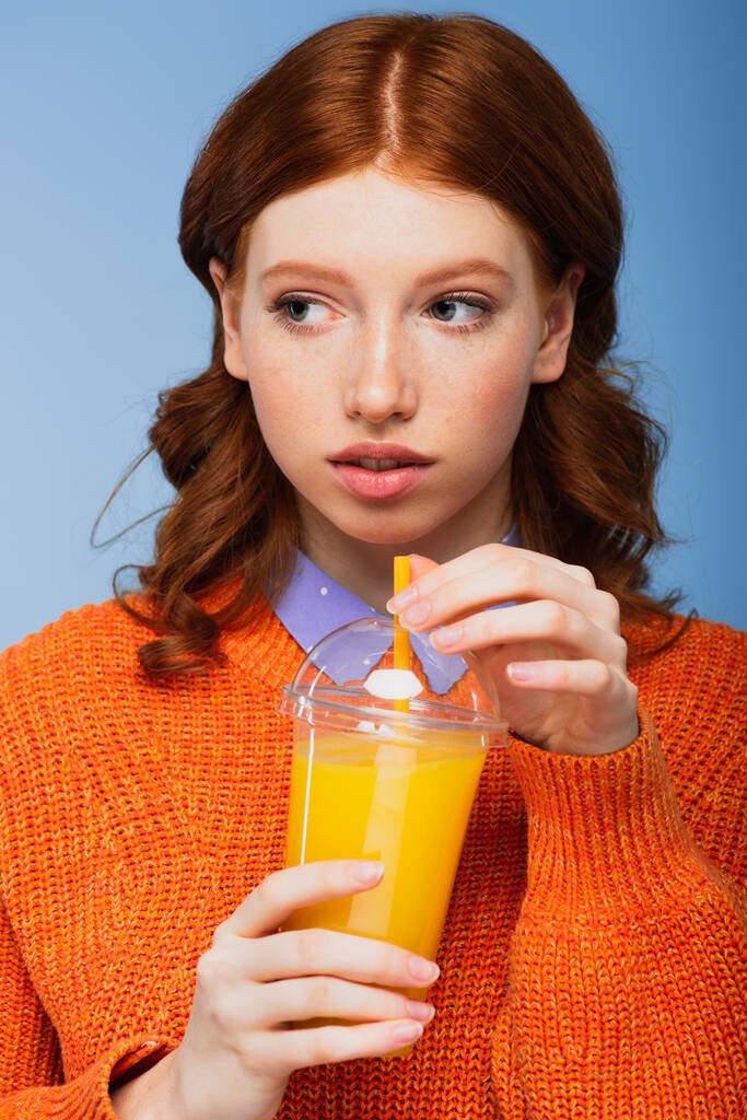 joven pelirroja en suéter sosteniendo taza de plástico fresco con jugo de naranja aislado en azul - Foto, imagen
