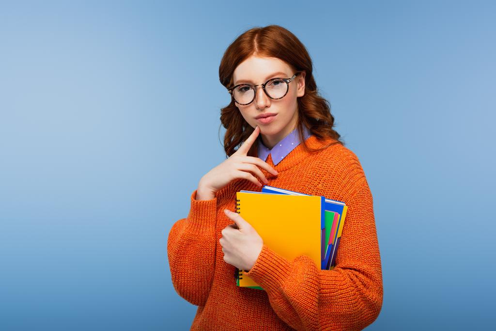 töprengő tanuló szemüvegben és narancssárga pulóverben, kék alapon izolált jegyzetfüzetekkel - Fotó, kép