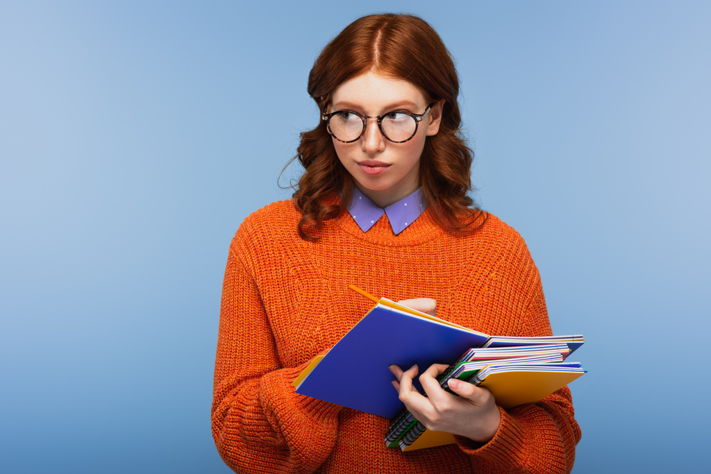 κοκκινομάλλα φοιτήτρια με γυαλιά και πορτοκαλί πουλόβερ κρατώντας σημειωματάρια και μολύβι που απομονώνονται σε μπλε - Φωτογραφία, εικόνα