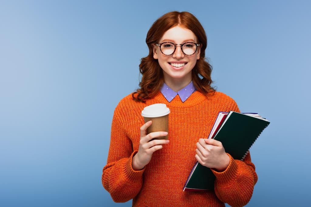 alegre joven estudiante en gafas y suéter naranja sosteniendo cuadernos y taza de papel aislado en azul - Foto, imagen