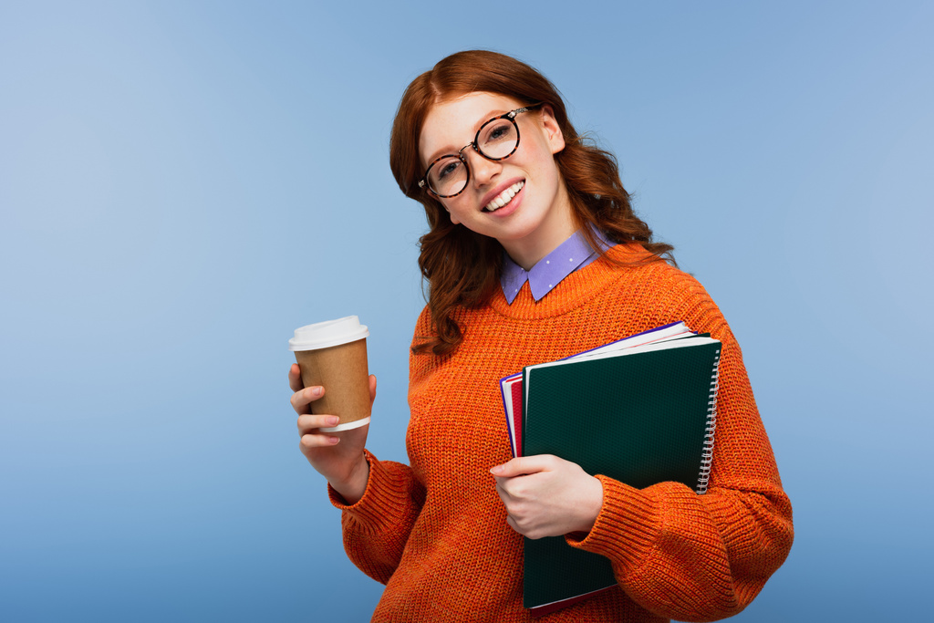šťastný zrzka student v brýlích a oranžový svetr drží notebooky a papírový pohár izolované na modré - Fotografie, Obrázek