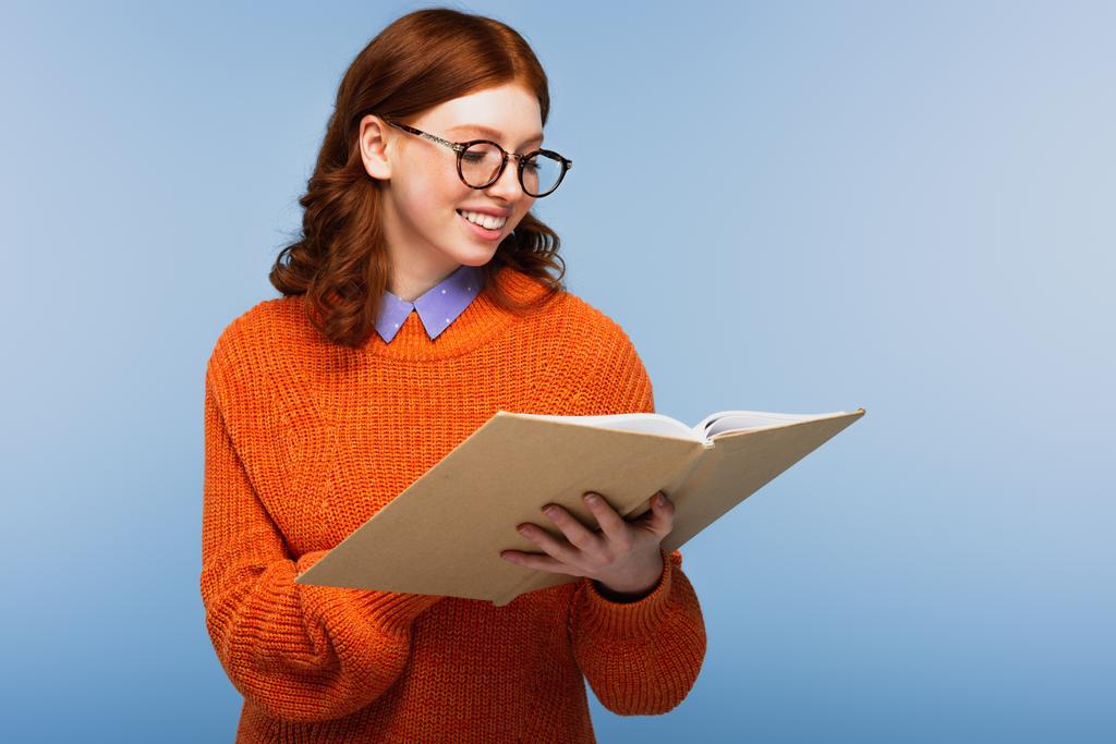 χαρούμενη κοκκινομάλλα μαθήτρια με γυαλιά και πορτοκαλί πουλόβερ βιβλίο ανάγνωσης απομονωμένο στο μπλε - Φωτογραφία, εικόνα