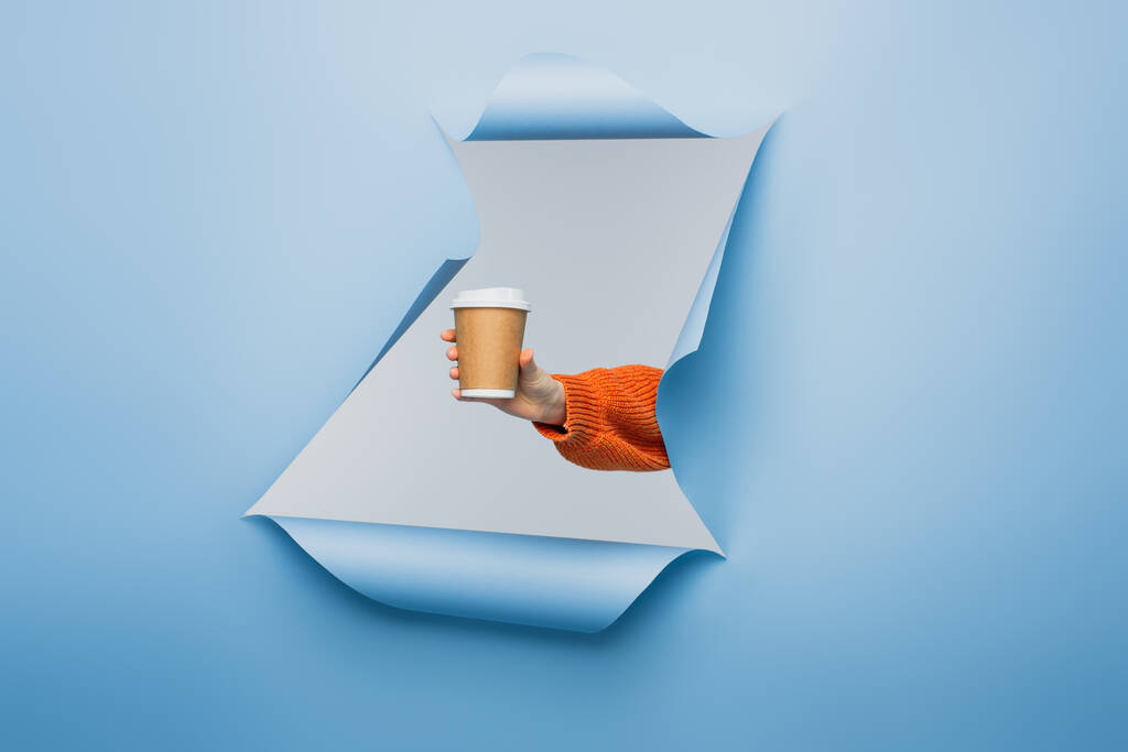 cropped view of young woman holding paper cup on blue ripped paper background  - Photo, Image