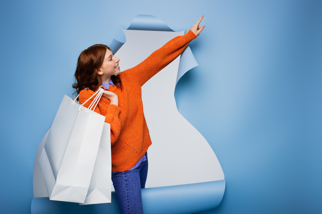 junge glückliche Frau hält Einkaufstüten in der Hand und zeigt mit dem Finger auf blau gerissenen Papierhintergrund  - Foto, Bild