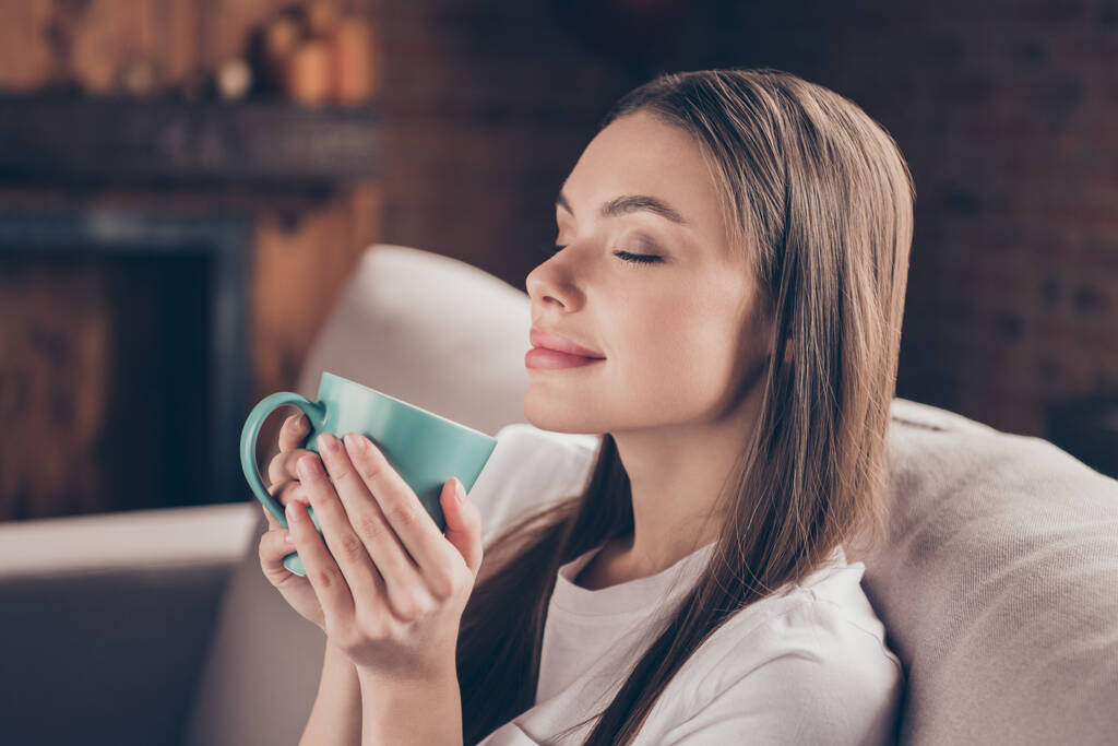 Kuva viehättävä suloinen nuori nainen pukeutua valkoinen t-paita istuu sohvalla juo teetä rentouttava suljetut silmät sisätiloissa talon huone - Valokuva, kuva