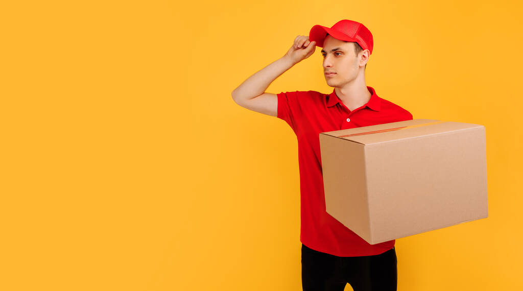 Happy excited male courier wearing red t-shirt and cap holding box parcel on isolated yellow background, express delivery, shopping concept - Photo, Image