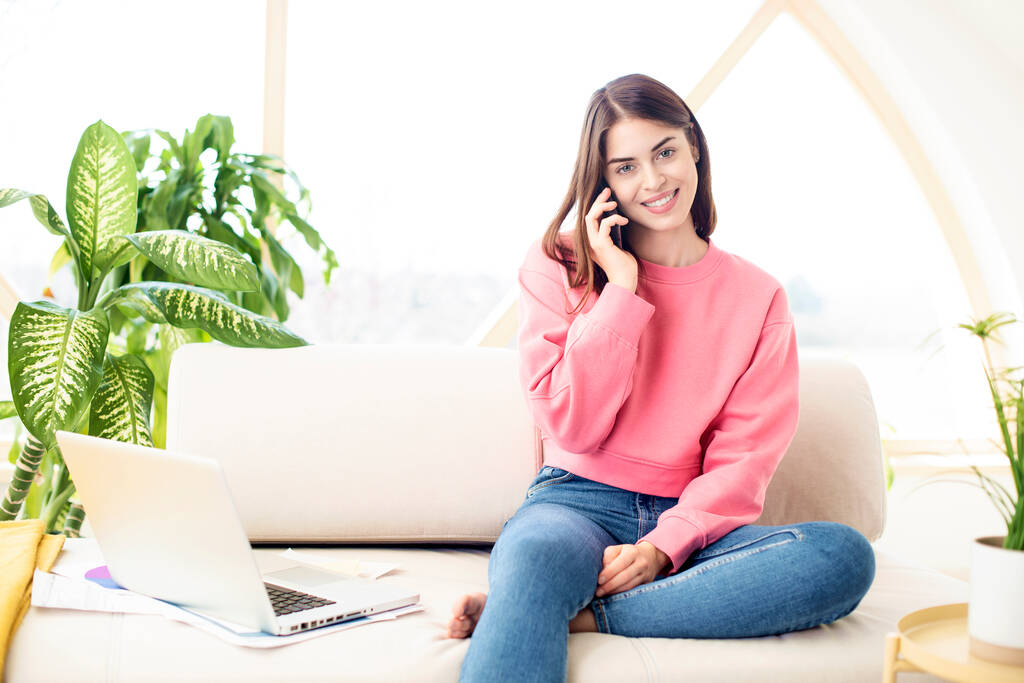 Aufnahme einer lächelnden jungen Geschäftsfrau, die mit jemandem auf ihrem Handy spricht und Laptop benutzt, während sie von zu Hause arbeitet. Innenbehörde. - Foto, Bild
