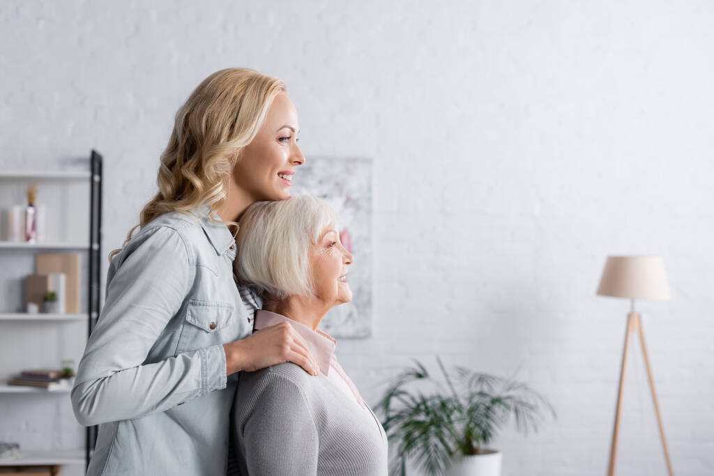 Side view of cheerful woman standing near senior parent in living room  - Photo, Image