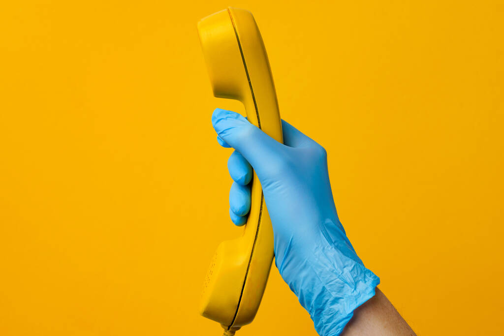 Γυναικείο χέρι σε ιατρικό γάντι κρατώντας κίτρινο τηλέφωνο ομιλητής - Φωτογραφία, εικόνα