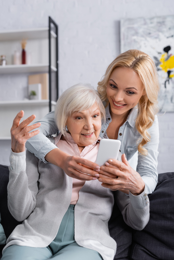 Usmívající se žena pomocí mobilu v blízkosti starší matky v obývacím pokoji  - Fotografie, Obrázek