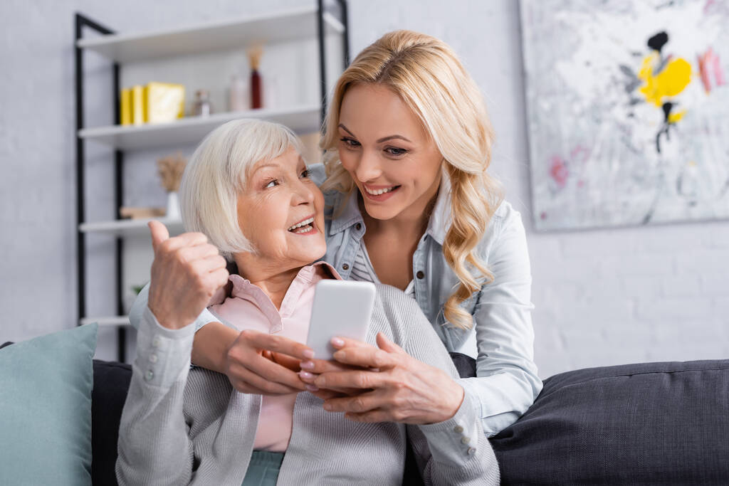 Χαρούμενη ηλικιωμένη γυναίκα κρατώντας smartphone και δείχνοντας σαν χειρονομία κοντά στην κόρη  - Φωτογραφία, εικόνα