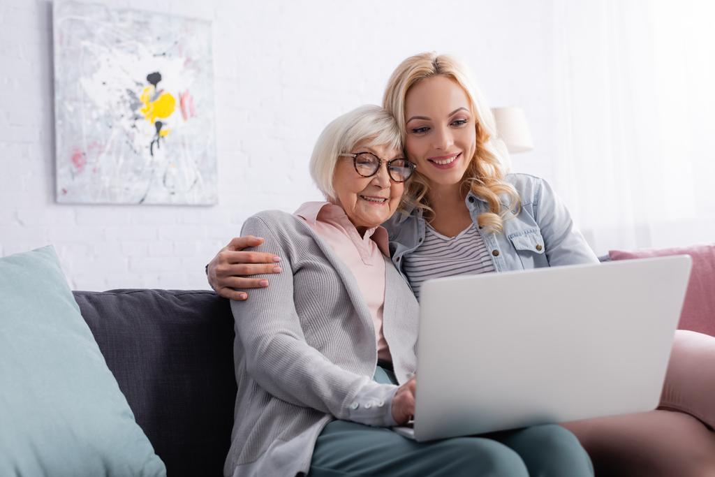 Усміхнена жінка обіймає старшу матір, використовуючи ноутбук у вітальні
  - Фото, зображення