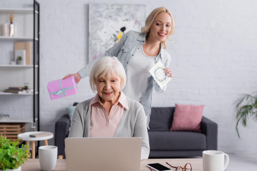 Senior vrouw met laptop in de buurt dochter met huidige en wenskaart op wazige achtergrond  - Foto, afbeelding