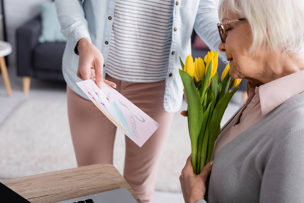 Senior woman holding tulips near daughter with greeting card at home  - Photo, Image