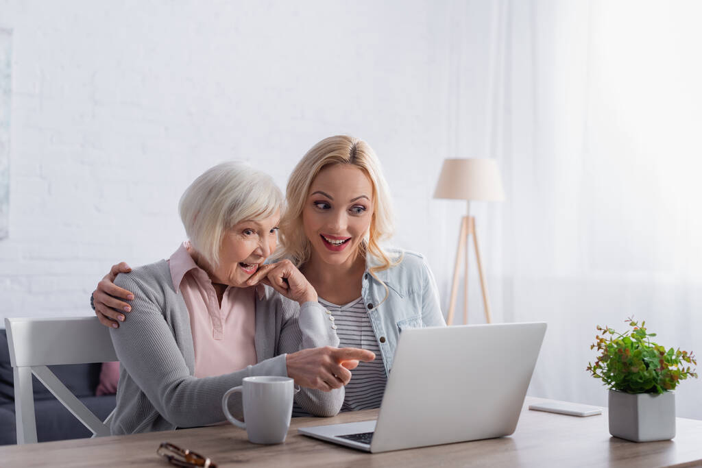 Fröhliche ältere Frau zeigt neben Tasse und Tochter auf Laptop  - Foto, Bild