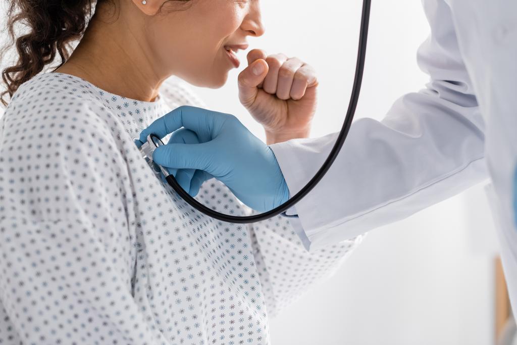 vista parcial del médico en guantes de látex examinando la tos mujer afroamericana con estetoscopio - Foto, imagen