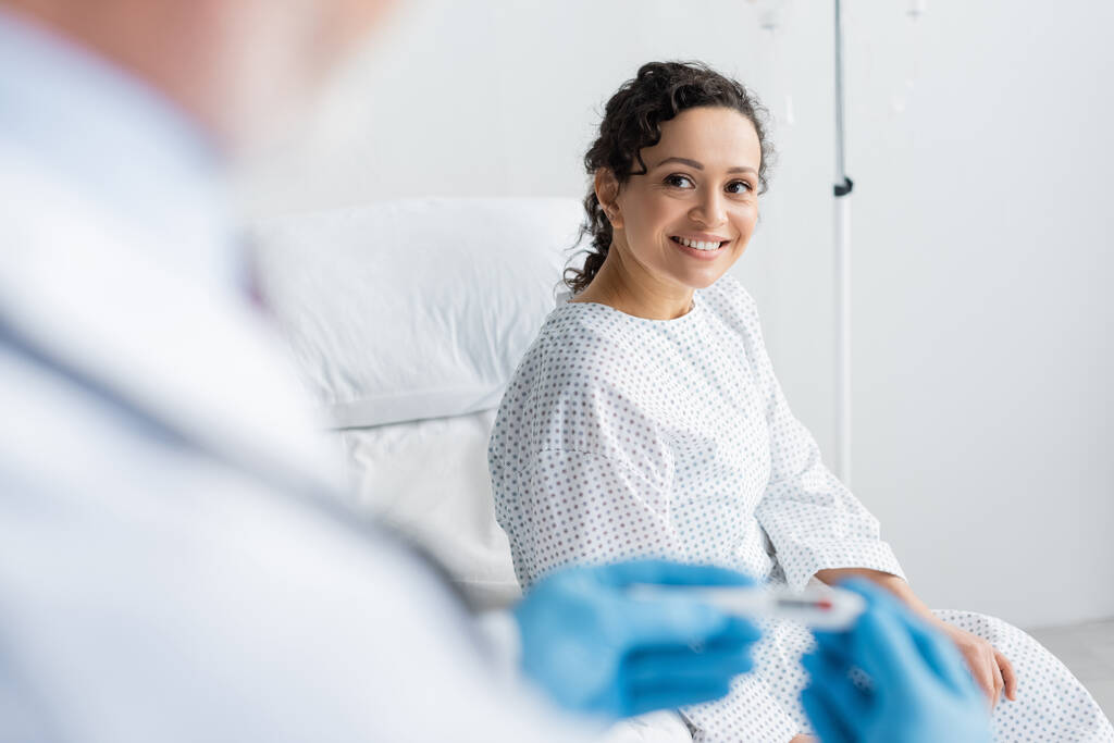 mulher afro-americana sorrindo perto de médico em luvas de látex segurando termômetro em primeiro plano borrado - Foto, Imagem