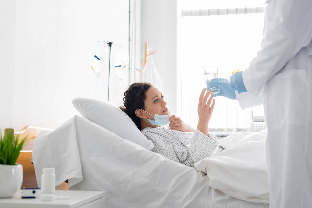 Arzt in Latexhandschuhen gibt kranker Afroamerikanerin im Krankenhausbett ein Glas Wasser - Foto, Bild