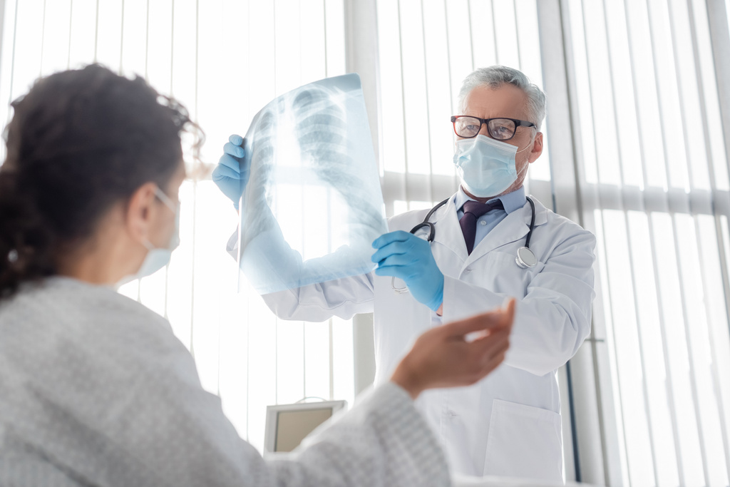 рентгенолог в медицинской маске держит рентген легких рядом с африканской американкой на размытом переднем плане - Фото, изображение