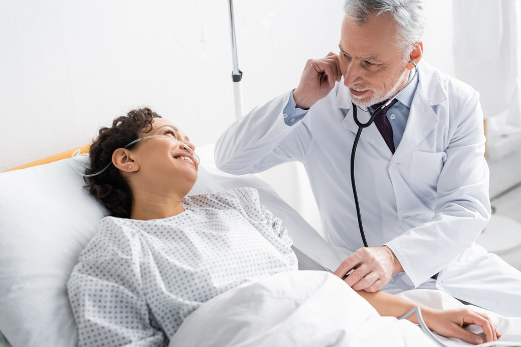 médico examinando mujer afroamericana sonriendo en cama de hospital - Foto, imagen