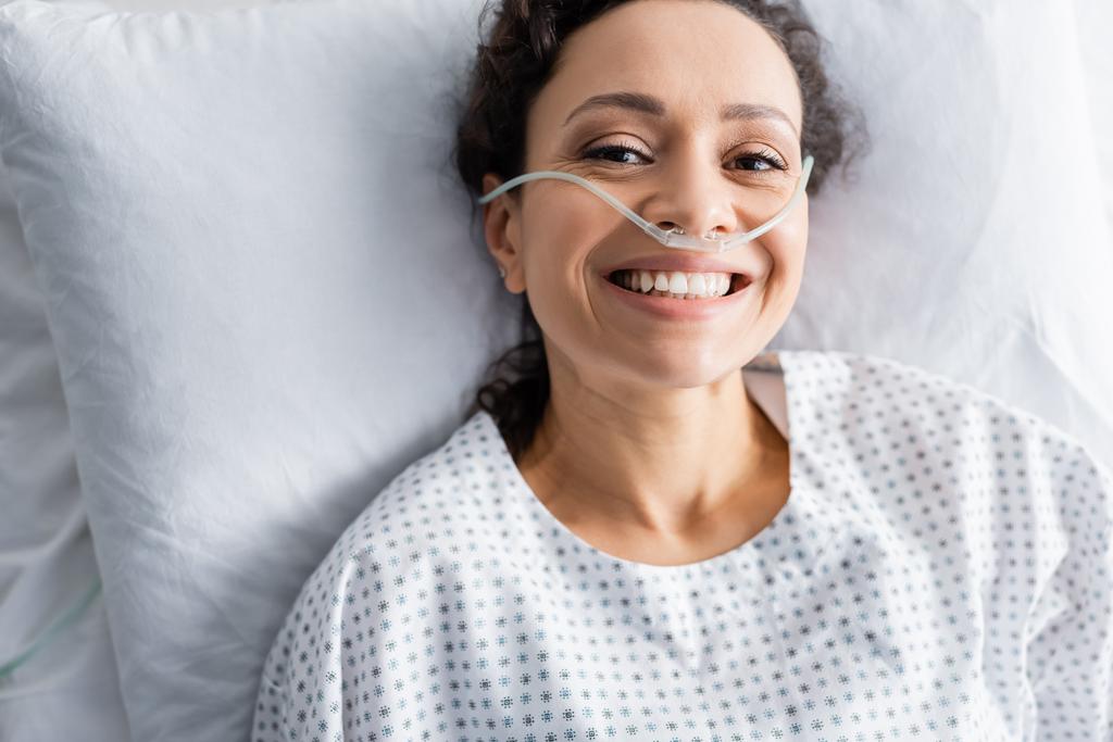 κορυφαία άποψη του χαμογελαστού Αφροαμερικανή γυναίκα με ρινική κάνουλα ξαπλωμένη στο κρεβάτι του νοσοκομείου - Φωτογραφία, εικόνα