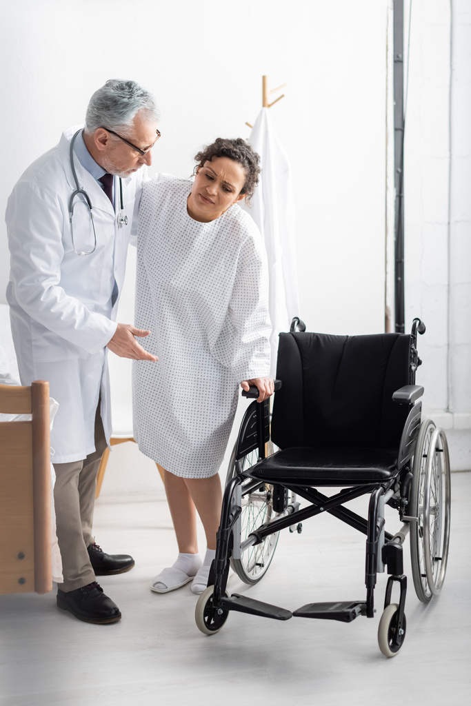 doctor apuntando a silla de ruedas mientras apoya a mujer afroamericana en hospital - Foto, Imagen