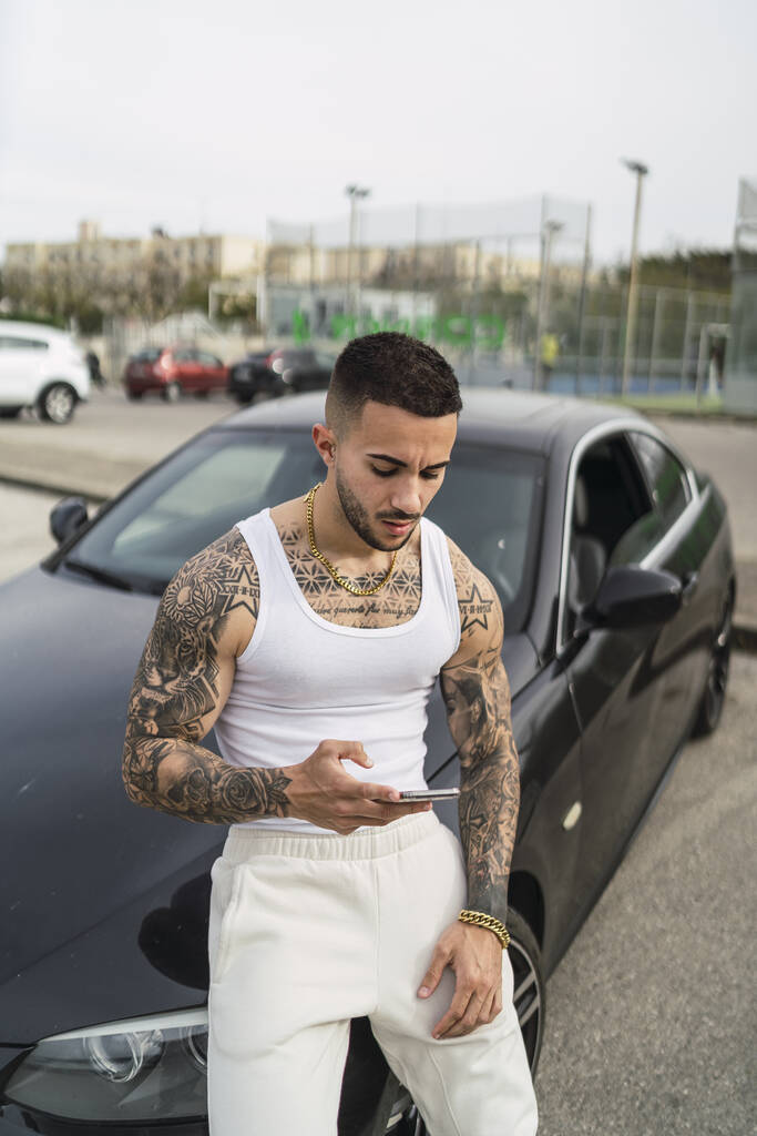Una toma vertical de un sexy hombre tatuado usando su teléfono apoyado en su coche - Foto, imagen
