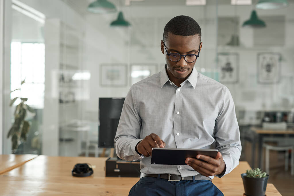 Joven empresario africano apoyado en su escritorio en una gran oficina moderna que trabaja en línea con una tableta digital - Foto, imagen