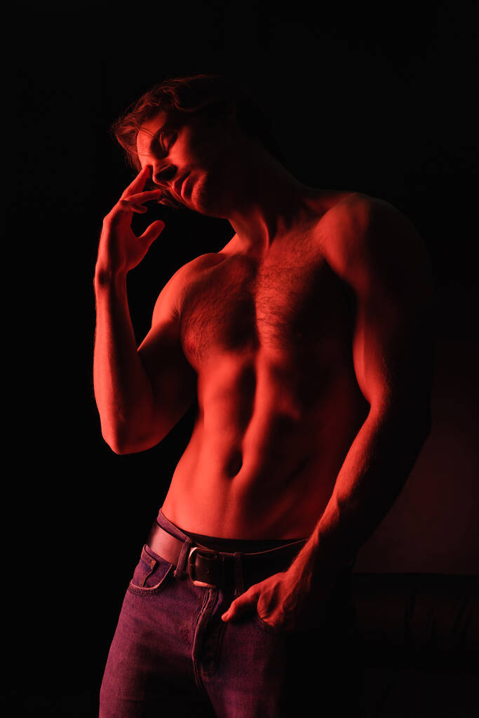 czerwone oświetlenie na ciele sexy muskularny mężczyzna stojący z ręką w kieszeni odizolowany na czarny  - Zdjęcie, obraz
