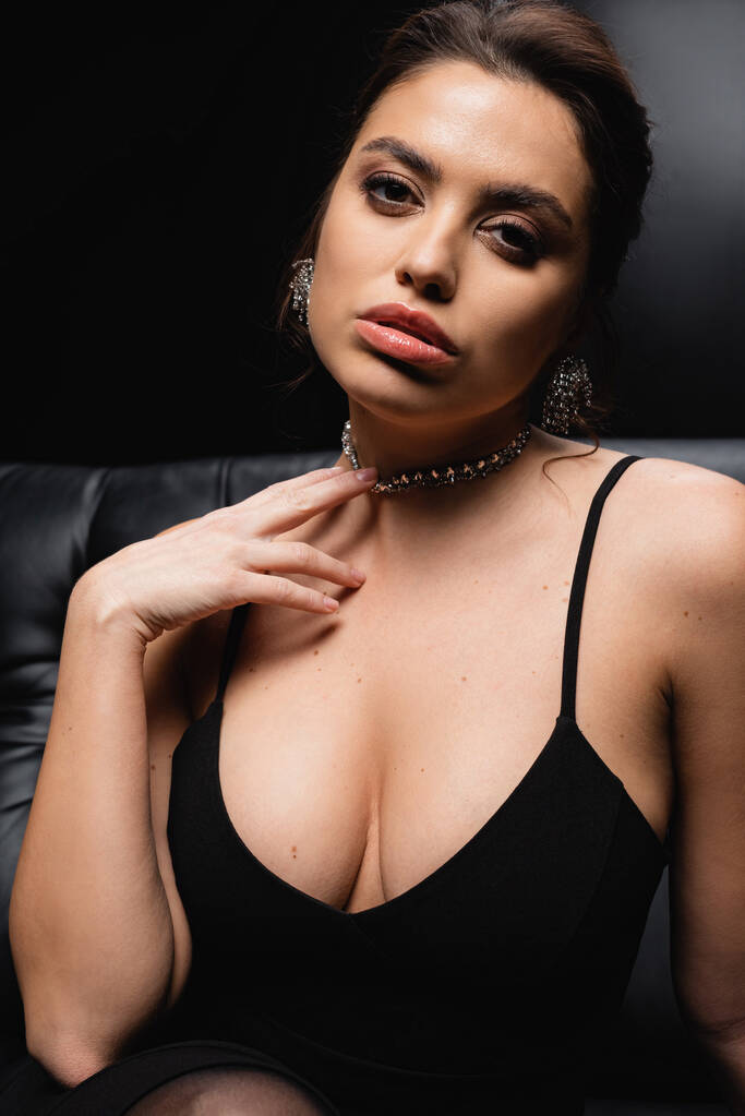 seductive woman in slip dress adjusting necklace and looking at camera on black - Photo, Image
